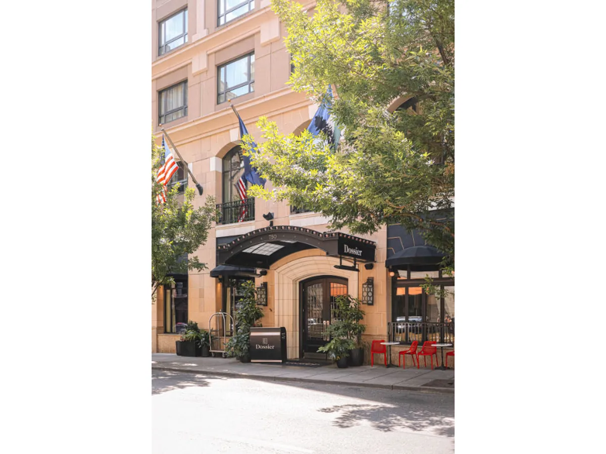 An inviting exterior scene with the hotel’s entrance archway, decorative flags, lush planters, and red outdoor chairs. The area is bathed in natural light, suggesting a lively place for guests to relax outdoors, socialize, or start their Portland adventure.