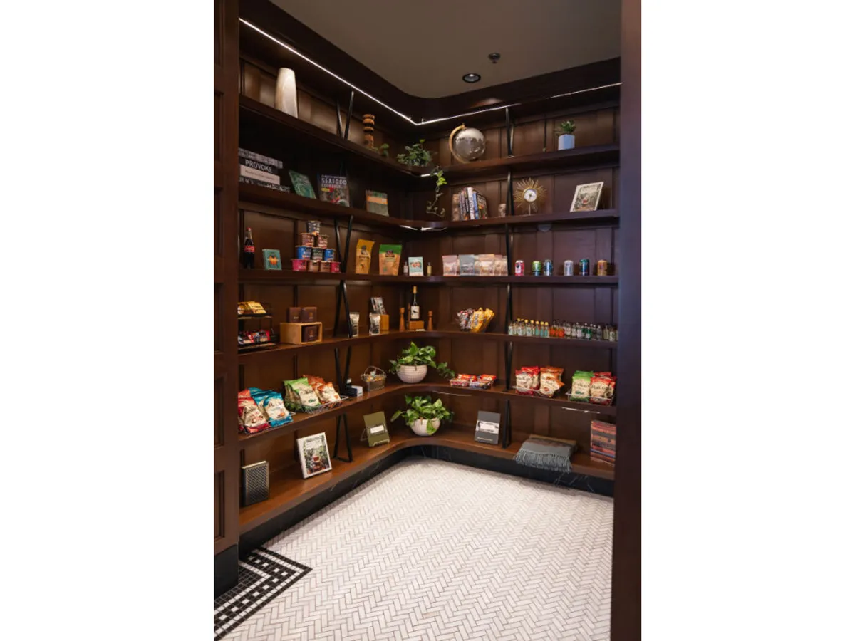 This image presents wooden shelves stocked with a variety of snacks and drinks, surrounded by lush green plants and decorative items. The scene feels cozy, inviting, and features guests casually browsing the offerings.