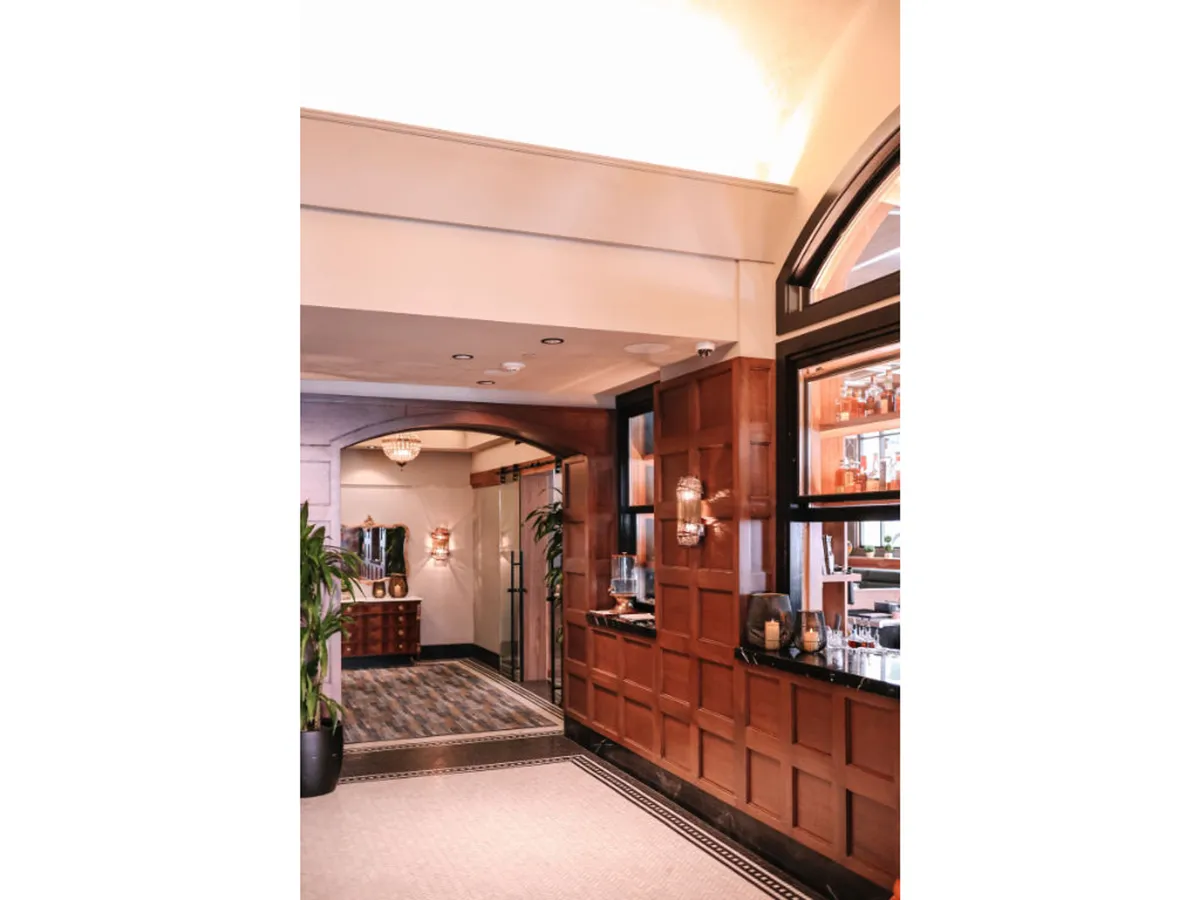 A warmly lit interior featuring elegant wood paneling, a chandelier, decorative lighting, and a bar area with plants. The space looks comfortable and sophisticated, fostering a welcoming atmosphere where guests can relax or seek assistance from attentive staff.