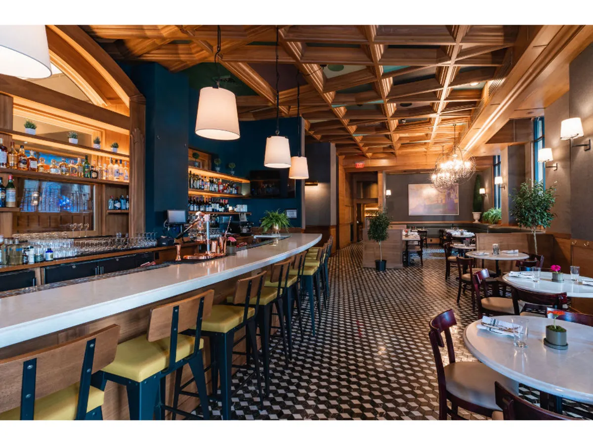 This image showcases a stylish bar area featuring a sophisticated counter, elegant chandeliers, and a decorative ceiling. The seating arrangement invites guests to relax and socialize, while the overall ambiance is refined yet approachable—perfect for enjoying inventive cocktails or small plates.