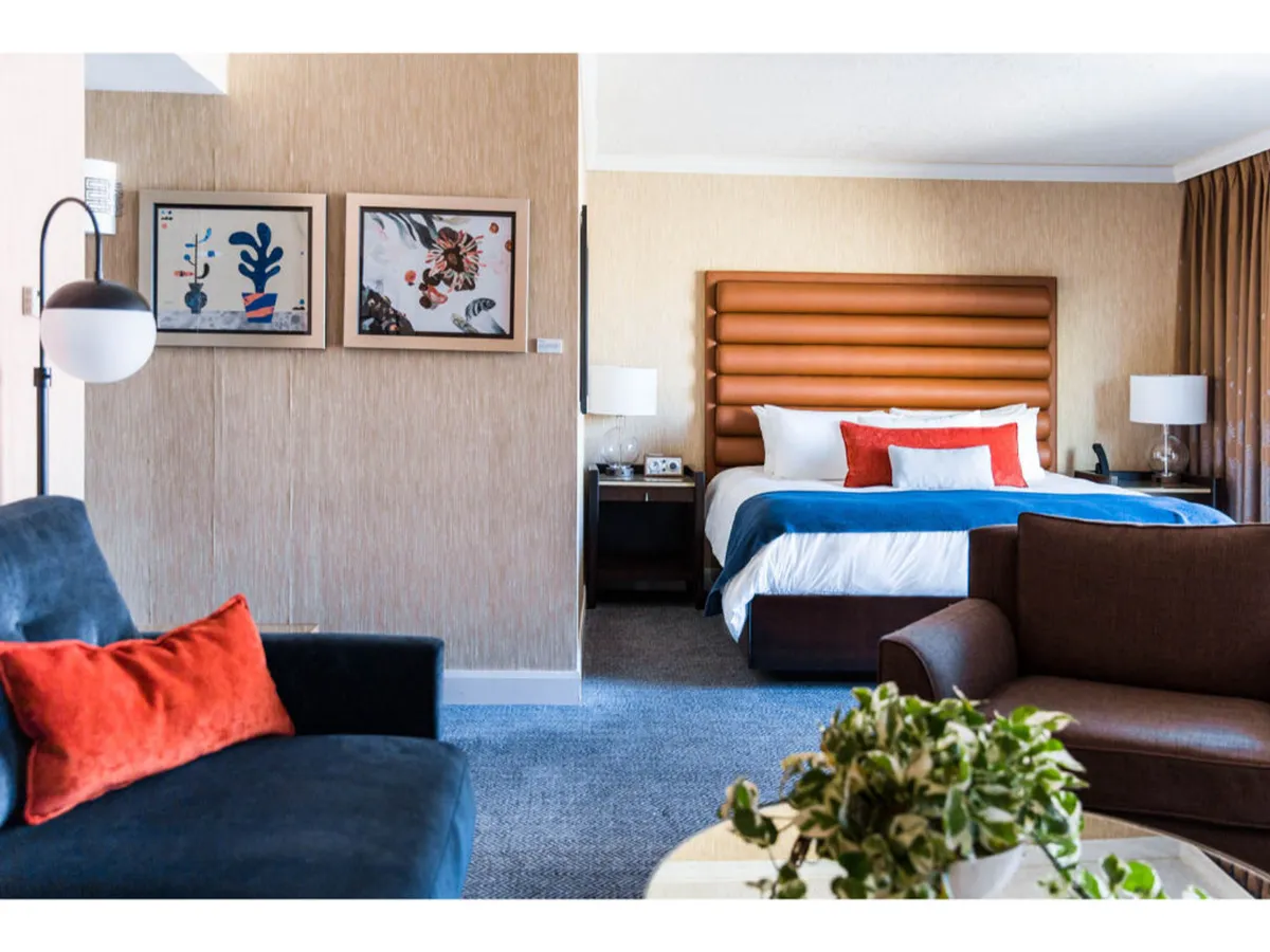 The image showcases a stylish hotel room with a large, inviting bed featuring a distinctive headboard, vibrant artwork above the bed, colorful cushions accenting a modern sofa, and a lush indoor plant. The overall ambiance is both cozy and contemporary, ideal for unwinding after a day exploring Portland's dynamic cityscape.