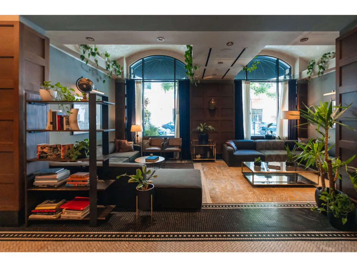 A suite filled with natural light from large windows, featuring comfortable seating areas, a decorative bookshelf, plants, and a coffee table. The arrangement showcases a relaxing yet functional space suited for reading, socializing, or working, with a clear sense of comfort and home-like atmosphere.