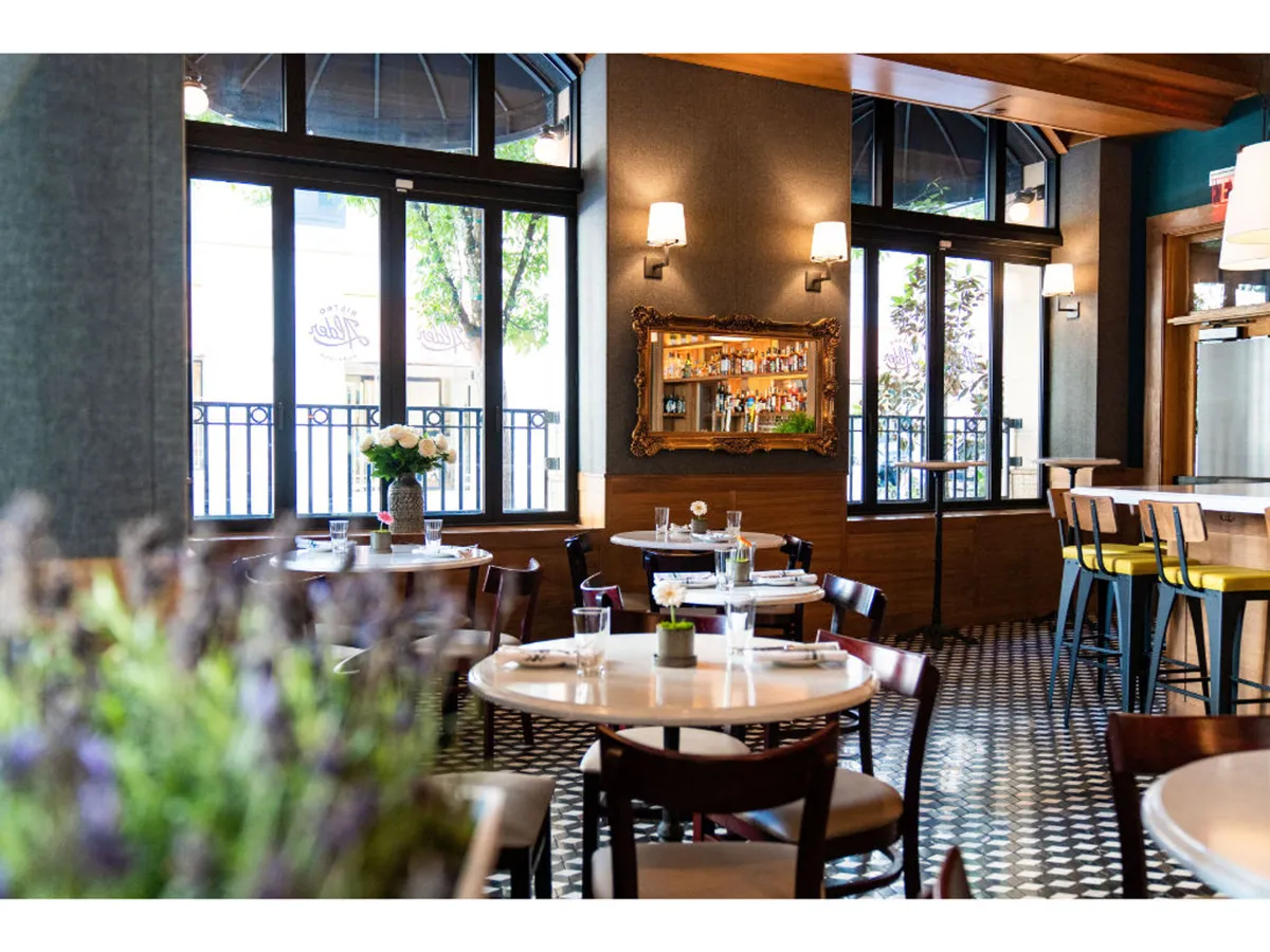 A cheerful, relaxed interior scene showing tables with fresh flowers, a well-stocked bar, and large windows that let in abundant natural light. The atmosphere feels welcoming and social, likely reflecting a dog-friendly cafe or patio where guests can unwind after exploring the city with their pets.